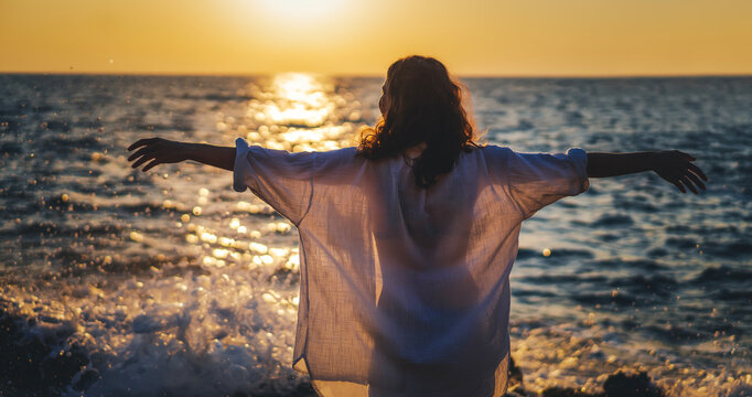 Young woman in a white shirt greeting the sun on the seashore at sunrise or sunset, with open arms