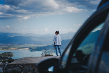 Woman traveler standing with stunning view of lake in mountains while travelling by car. Auto travel concept