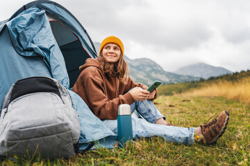 Naklejka na ściany i meble Young cheerful woman traveling with a tent using a smartphone. Hiking and trekking lifestyle