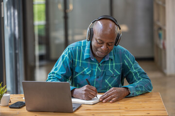 Senior black man wearing headphones enjoying listening to music or something interesting to relax while working. Study and take notes of interesting and useful things. - obrazy, fototapety, plakaty