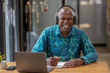 Senior black man wearing headphones enjoying listening to music or something interesting to relax while working. Study and take notes of interesting and useful things.