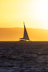 Yacht sailing in the Aegean sea in front of mountainous island filled by vivid orange sunset light