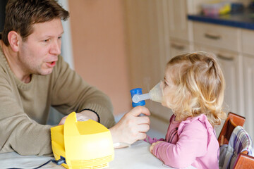 Little toddler girl making inhalation with nebulizer at home. Father helping and holding the...