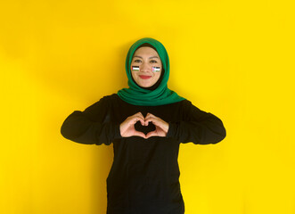 Beautiful young woman in hijab and Palestinian flag on her cheek making heart gesture with finger 