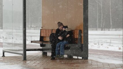Happy mother hugs son and smiles while sitting on bench on winter snowy day, wide shot. Family at wooden eco public transport stop, sheltered from snowfall, awaits arrival of bus.