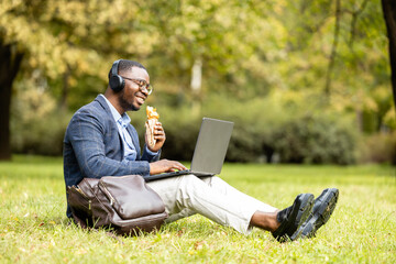 Businessman working outside the office. Sitting on grass in the park, eating his lunch and working on laptop.