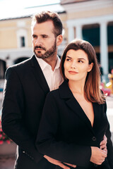 Beautiful fashion woman and her handsome elegant boyfriend in black suit. Sexy brunette model in jacket. Fashionable confident couple posing in street. Brutal man and female outdoors