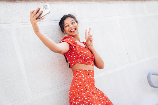 Young beautiful smiling hipster woman in trendy summer red dress. Carefree woman posing in the street near wall. Positive model outdoors in at sunny day. Cheerful and happy, takes selfie photos