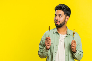 Ready to eat. Hungry Indian man waiting for serving dinner dishes with with restlessness holding cutlery fork knife will appreciate delicious restaurant meal. Excited guy isolated on yellow background - obrazy, fototapety, plakaty