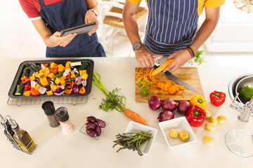 Diverse couple chopping vegetables and using tablet in sunny kitchen at home
