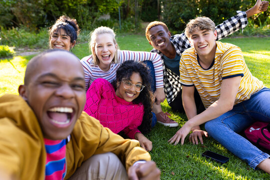 Portrait of happy diverse teenage friends taking selfie and sitting on grass in park