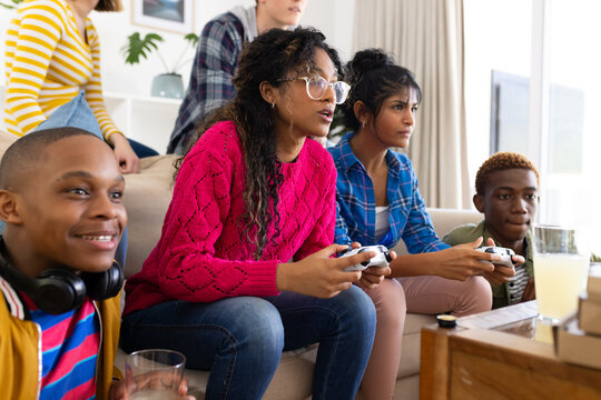 Happy diverse group of teenage friends sitting on couch and playing video games with snacks at home