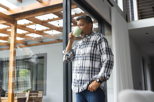 Thoughtful senior biracial man drinking coffee at window at home