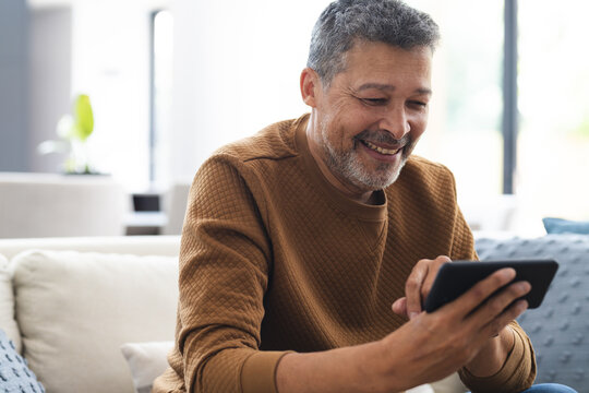 Happy senior biracial man sitting on couch and using smartphone at home
