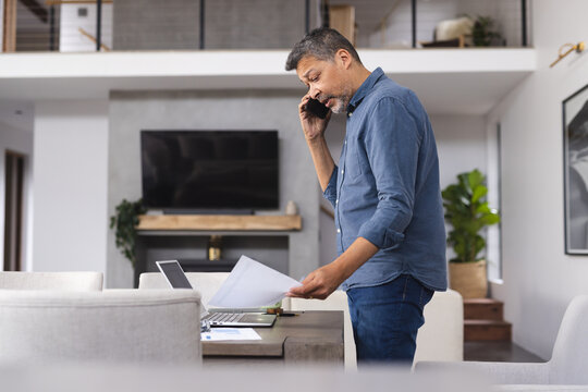 Busy senior biracial man talking on smartphone and working on laptop at home