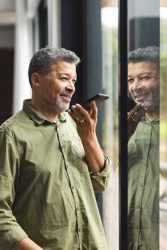 Happy senior biracial man talking on smartphone at window at home