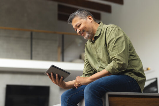Happy senior biracial man sitting on table and using tablet at home