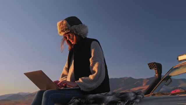 Young Girl hiker sits on trunk a car, uses a laptop, works. Remote freelance work in the mountains. The concept of remote learning, freelancing and modern technology. Outdoor adventure lifestyle.