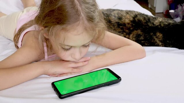 A child looks at a green phone screen in bed with a black cat. Chromed green screen. Communication, game or cartoon on the phone