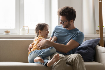 Loving cheerful young father sit on cozy sofa with little daughter at home, play, spend time together. Cute girl tickling laughing handsome father, have fun in living room. Relationship, fatherhood