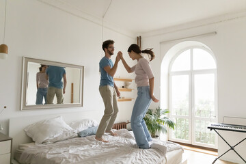 Young millennial carefree couple spend free time at home jumping on bed in cozy bedroom, have fun, enjoy family weekend at modern apartment, celebrating purchase of new house. Date, relationship, mood - obrazy, fototapety, plakaty