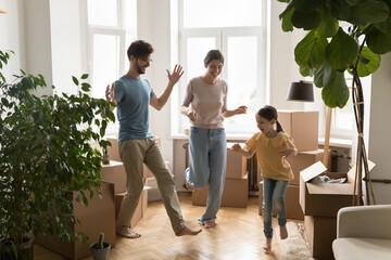 Happy active parents and cute little girl dancing to energetic music and jumping together among boxes in new apartment. Overjoyed homeowners celebrate moving at new house, have fun with cute daughter