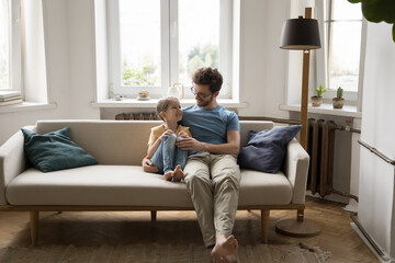 Happy handsome father sit on sofa hug little cute daughter, talk to child, enjoy communication,...