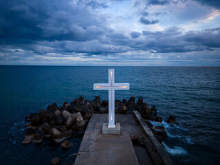 A large Christian cross stands at the edge of a pier against a dramatic sky and sea, seen from above