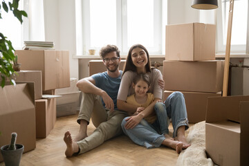 Cheerful young spouses with little cute daughter sit on floor near cardboard boxes with belongings,...