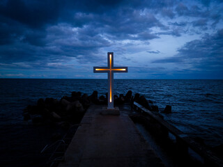 A large Christian cross stands at the edge of a pier against a dramatic sky and sea, seen from above
