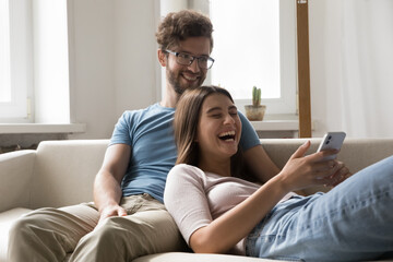 Joyful attractive young spouses relaxing on cozy sofa, using cellphone watch funny videos on smartphone, spend weekend time at home, having fun with new mobile application, enjoy internet amusement