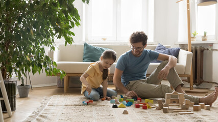 Cheerful daddy and cute little daughter play toy blocks on heating floor, communicate, have fun in...