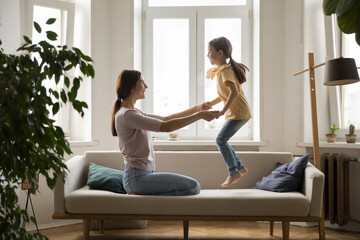 Cheerful mother play with little cute daughter on couch, mom holding hands while joyful girl...