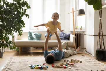 Little girl enjoy playtime with dad at home. In living room young man lying on floor lifts his adorable daughter on outstretched arms, kid pretend flying on air stretched her hands like plane wings - obrazy, fototapety, plakaty