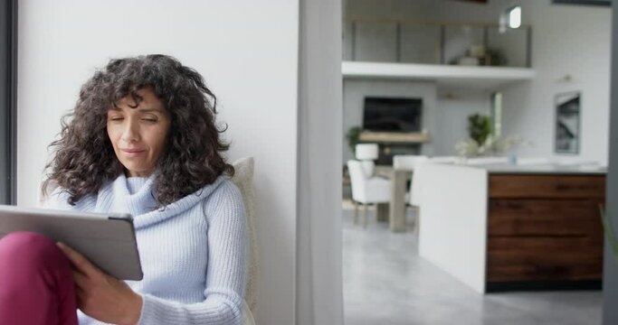 Happy mature caucasian woman sitting at window and using tablet at home, slow motion