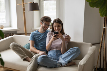 Happy couple relax together on sofa in living room with mobile phone, look at screen device, share funny content in social networks, have fun use modern technology. E-services, internet, social media