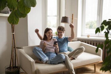 Joyful young couple use mobile phone together relaxing on sofa at home, looking at cellphone screen feel happy read email about bank loan acceptance, celebrate online lottery win. Modern tech, victory - obrazy, fototapety, plakaty