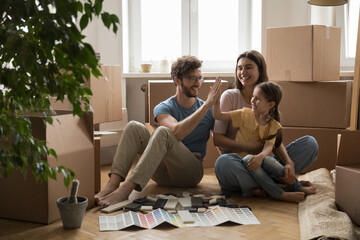 Happy young couple with little 6s daughter sit on floor with cardboard boxes and heap of colorful...