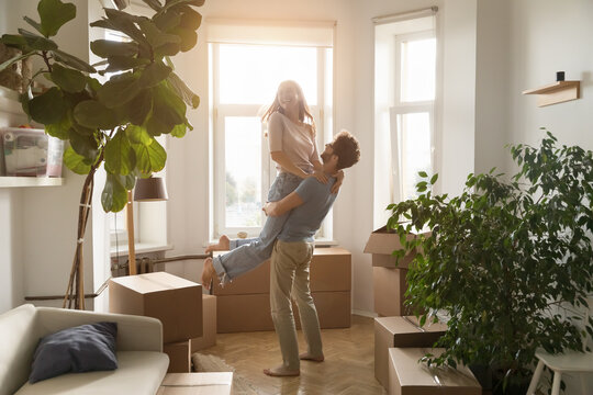 Happy husband lifts his young beautiful wife, laughing, swirling, enjoy move-in day to new, first house, big carton boxes nearby in unfurnished sunny living room. Bank loan for young family, new life