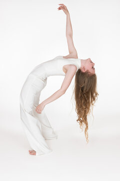 Full length view of flexible young woman dancer bending over backwards with eyes closed. Side view of slim barefoot model with long wavy hair dressed in white jumpsuit. Studio shot on white background
