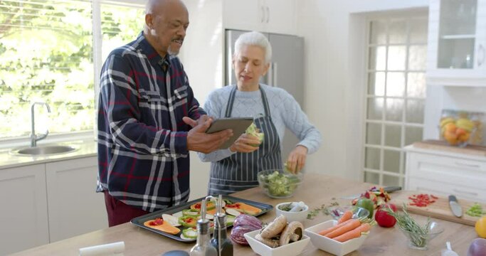 Happy diverse senior couple preparing vegetables, using tablet in kitchen, copy space, slow motion