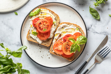 Two sandwiches or toasts with tomatoes, cream cheese, olive oil and basil on a plate on white...