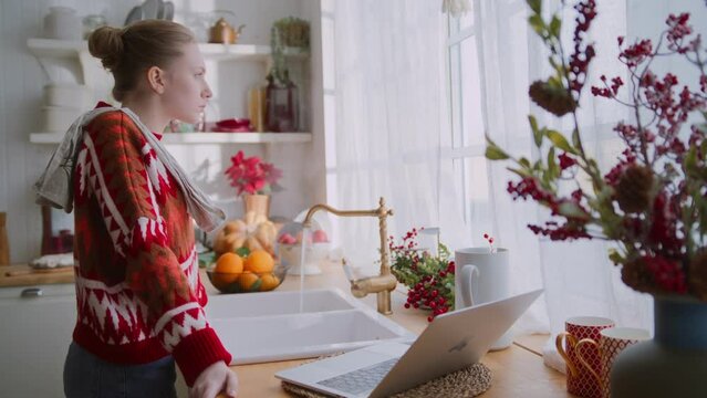 young woman in red sweater stays in kitchen leaning on sink feeling upset. thoughtful and frustrated girl at home during homework looks through window at christmas holidays