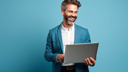 In formal attire, a businessman confidently gazes at a laptop with a friendly smile while standing against a blue studio background