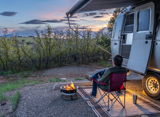 Matured Caucasian man, sitting on a chair with a cell phone by a camper warming by a campfire with light glowing behind and dark clouds overhead, McPhee Recreation Area, Colorado