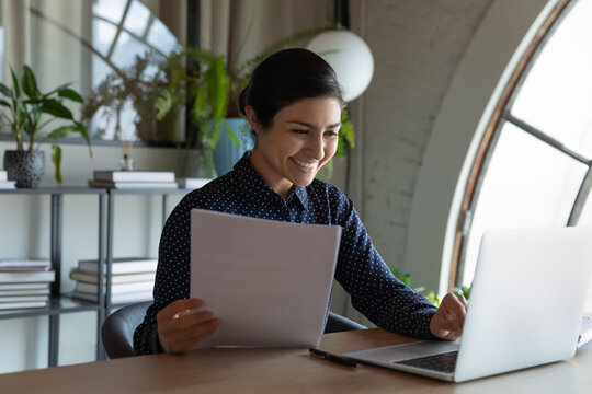 Happy Indian business woman, office employee reviewing paper contract, reading legal document, consulting Internet, working, using laptop. Satisfied professional checking report, doing paperwork