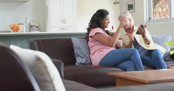 Happy diverse senior women using smartphone talking on sofa in sunny living room, slow motion