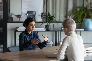 Indian female business mentor training intern, explaining work task to employee. Candidate and employer, recruiter, hr agent talking at job interview, meeting at table, discussing hiring, experience - obrazy, fototapety, plakaty