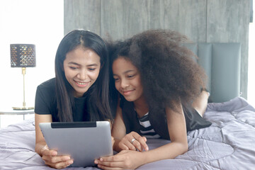 Happy family love bonding at home, African Asin mixed race daughter girls with curly hair and mother enjoy using digital table online surfing internet social media together, lying on bed in bedroom.