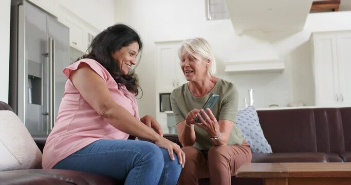 Happy diverse senior women using smartphone and discussing in sunny living room, slow motion